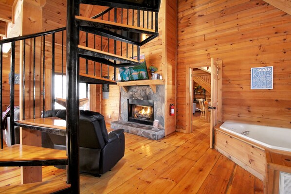 Een houten cabin interieur met een open haard, een stenen haardmantel, een zwarte trap, een houten tafel, een zwarte leren fauteuil en een bad.