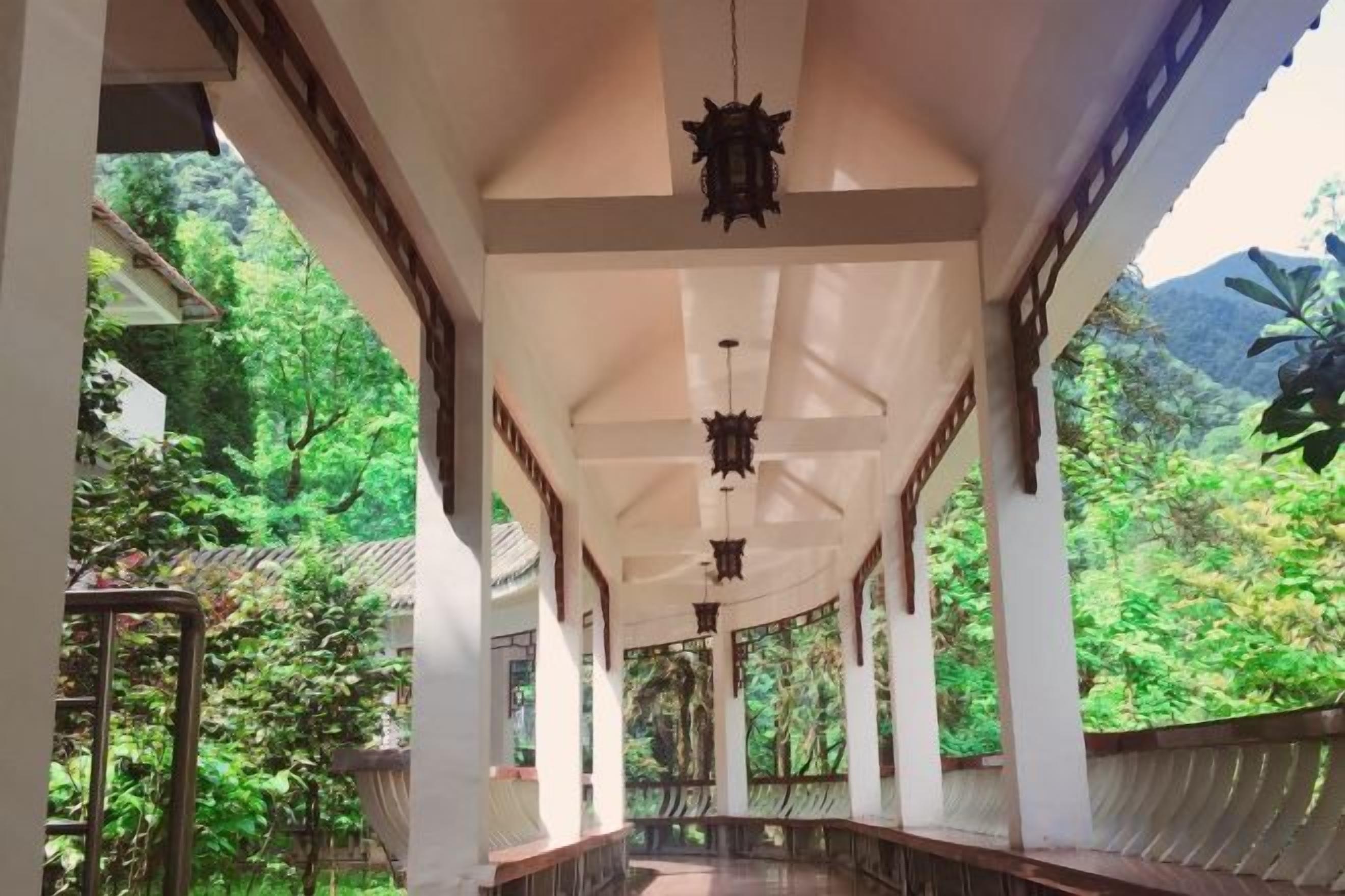 A covered walkway with columns and hanging lanterns, surrounded by lush greenery.