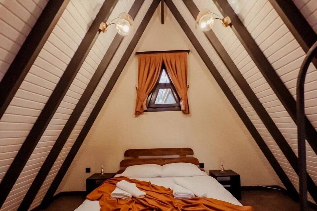 Um quarto de sótão aconchegante, com teto inclinado, uma pequena janela com cortinas, uma cama bem arrumada e duas luminárias de cabeceira.