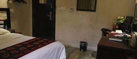 A hotel room with a bed, a desk, a chair, a trash bin, and a door.