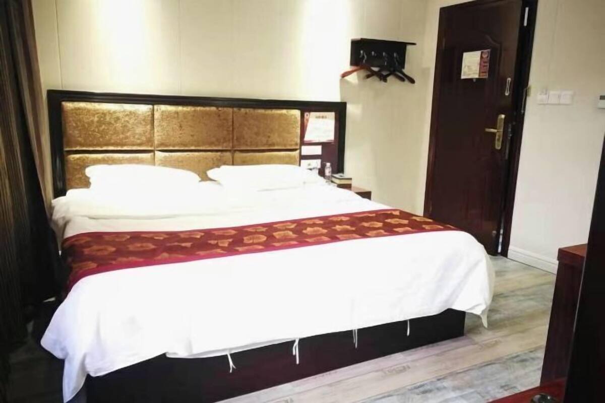 A hotel room with a large bed, a wooden headboard, a bedside table with a lamp, and a door with a coat rack.