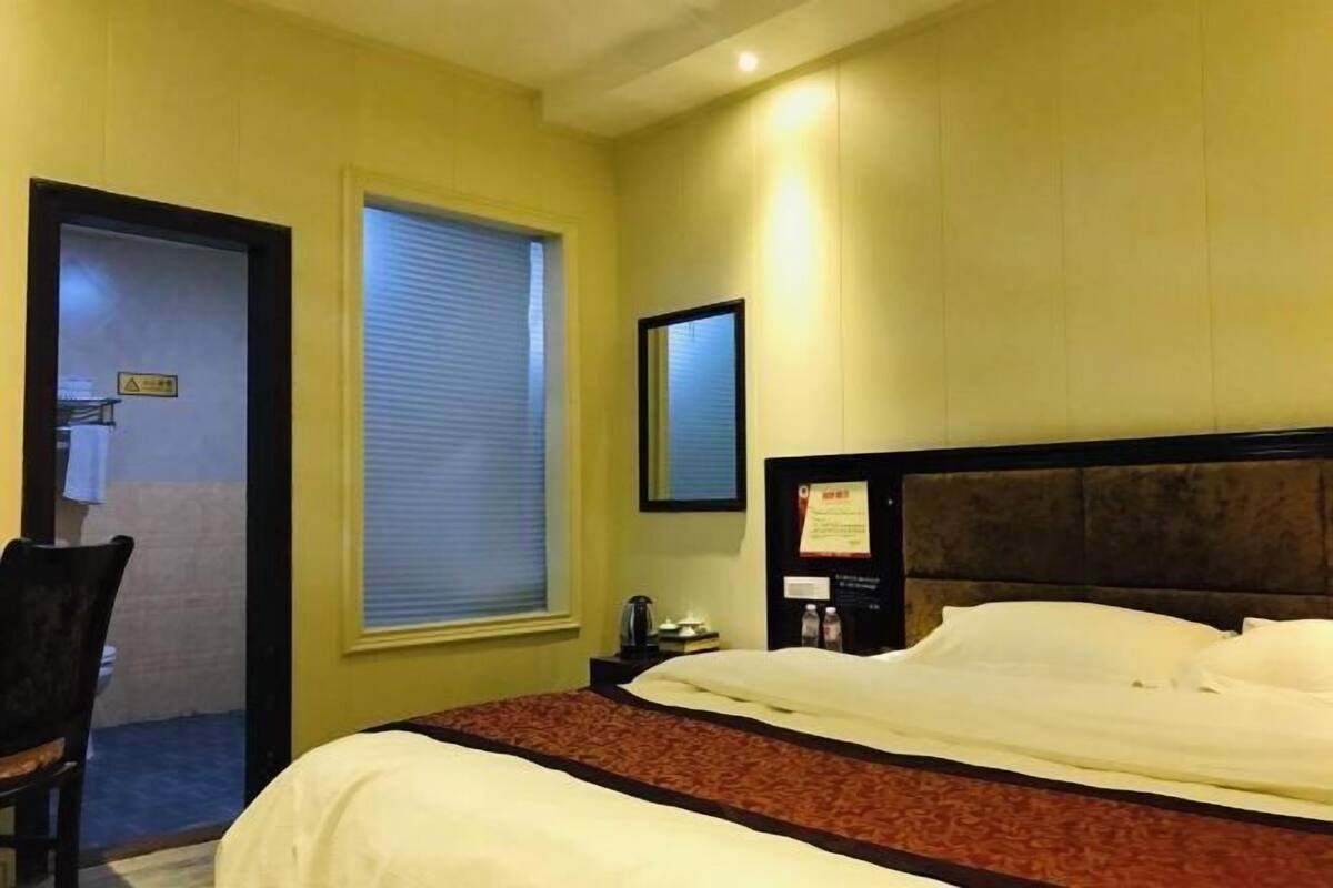 A hotel room with a bed, a chair, a window with blinds, a framed picture on the wall, and a nightstand with a lamp.