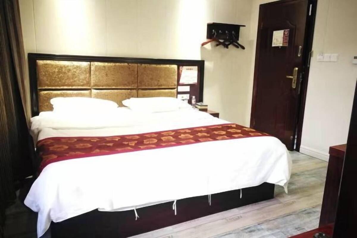 A hotel room with a large bed, a wooden headboard, a dark wooden door, and a small table with a lamp.