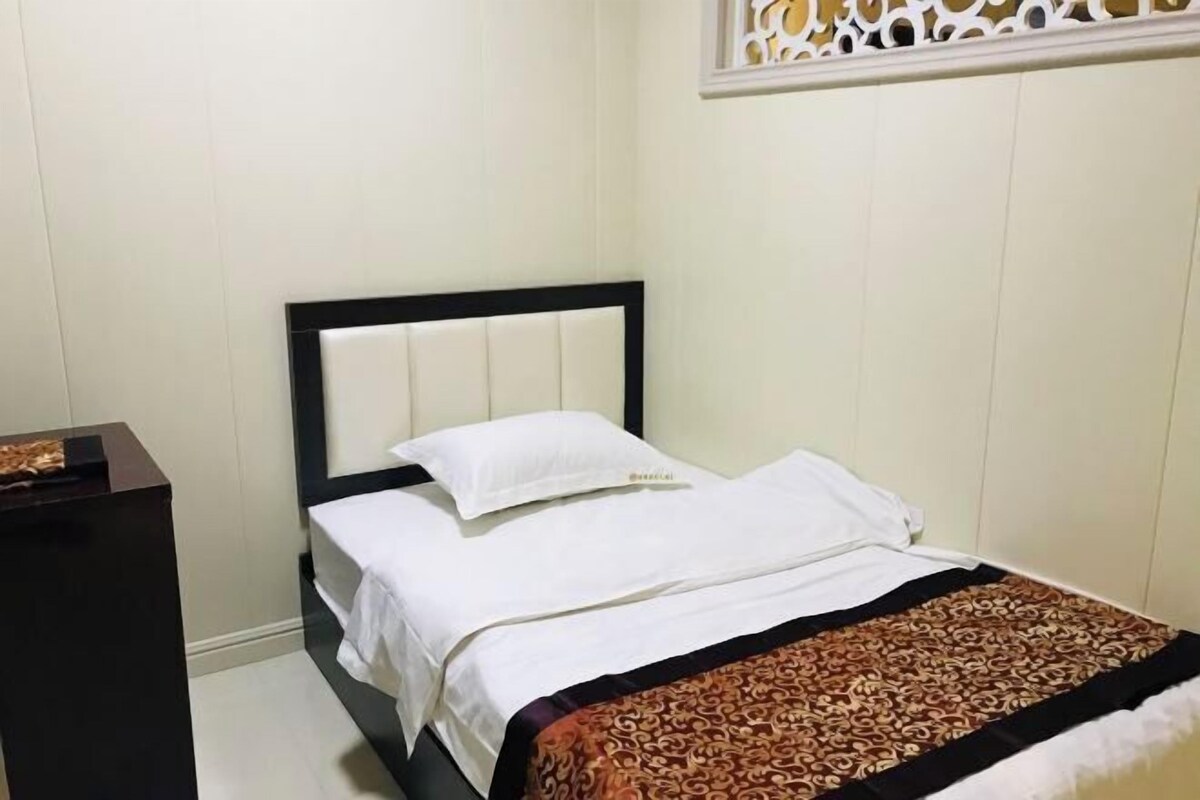 A neatly made bed with a white duvet and a patterned throw blanket, a dark wooden nightstand to the left, and a decorative wall panel above the bed.
