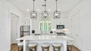A modern kitchen with a large island, bar stools, white cabinetry, and a stainless steel refrigerator. - Brand-new townhome with porch, W/D, golf course, pool, & beach access (Port Aransas)