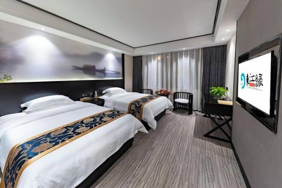 A hotel room with two beds, a desk, a chair, a TV, and a large window with curtains.