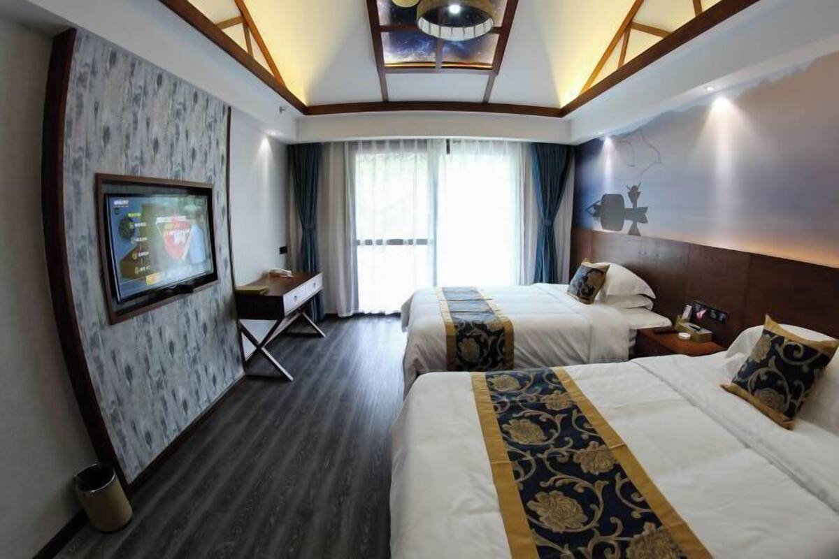 A hotel room with two beds, a desk, a television, and a large window with curtains.