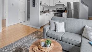 A modern living room with a grey sofa, a wooden coffee table, and a kitchen in the background. - Harbourview Loft B Two Bedroom Loft Apartment Located in Downtown Summerside (Summerside)