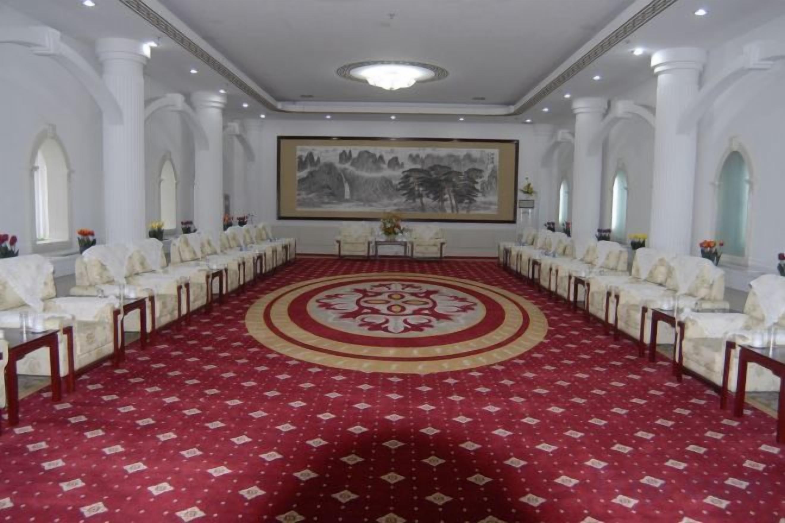 Un salón de conferencias con alfombra roja, sillas blancas y un cuadro grande en la pared.