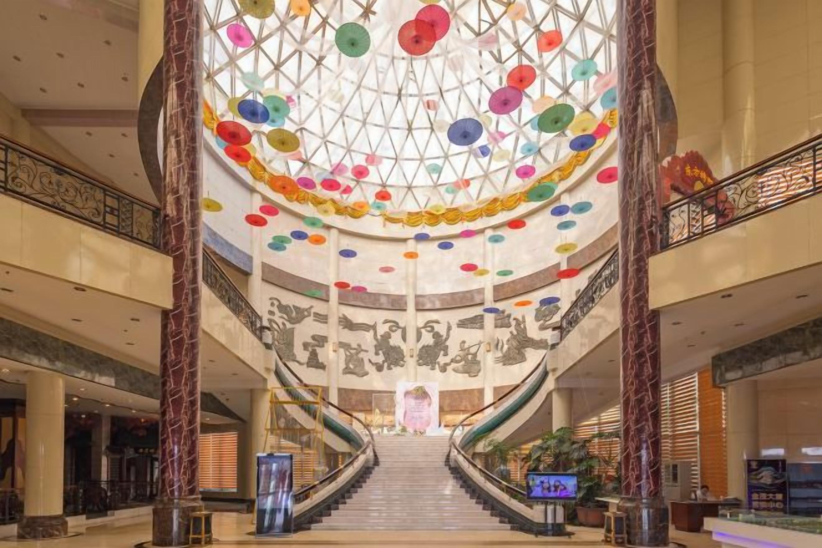 Un imponente atrio con una escalera central, techo de cristal con decoraciones circulares de colores y un gran mural en las paredes.