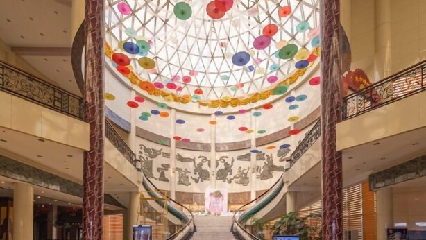 Um amplo átrio com escadaria central, colunas de mármore e teto decorado com detalhes circulares coloridos.