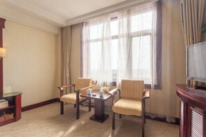 A hotel room with a large window, two armchairs, a small table with a teapot, and a television on a wooden stand.