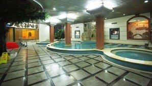 Uma grande piscina interna com várias pistas de natação, cercada por colunas de pedra e piso ladrilhado.