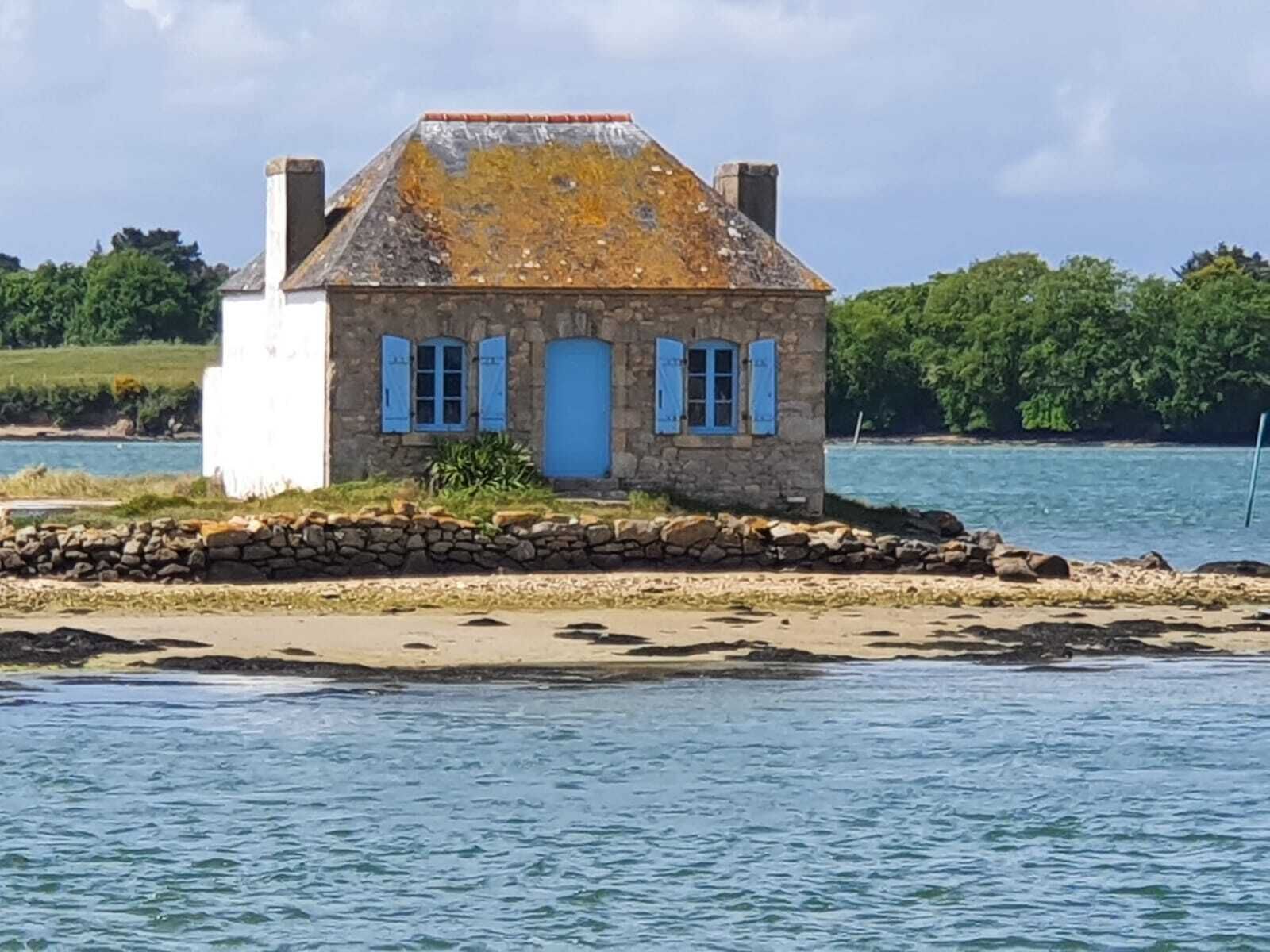 Ein aus Stein gebautes Haus mit blauer Tür und Fensterläden, gelegen an einer felsigen Küstenlinie mit einem Gewässer im Hintergrund.