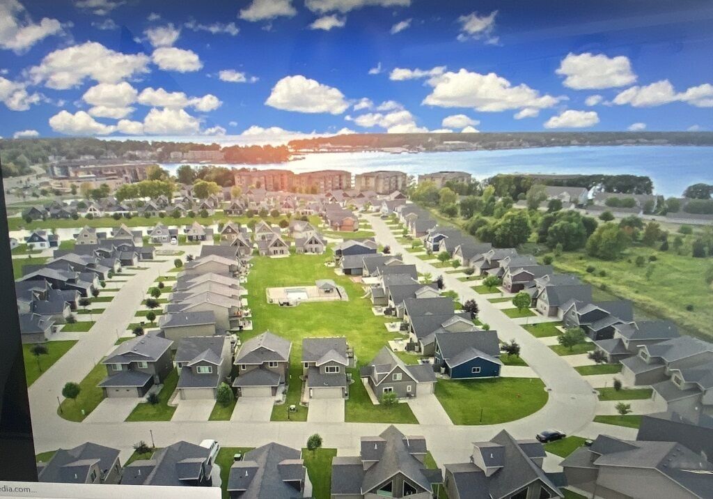 A residential area with houses arranged in rows, a central green space, and a body of water in the background.