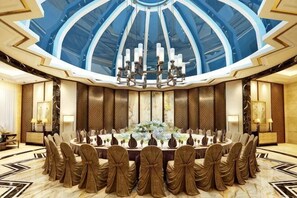 Un comedor formal con techo alto y un candelabro central, rodeado de mesas dispuestas para una comida.