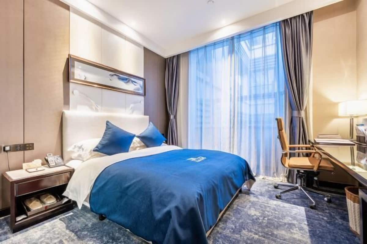 A hotel room with a large bed, a desk with a chair, a window with curtains, and a wall-mounted artwork.