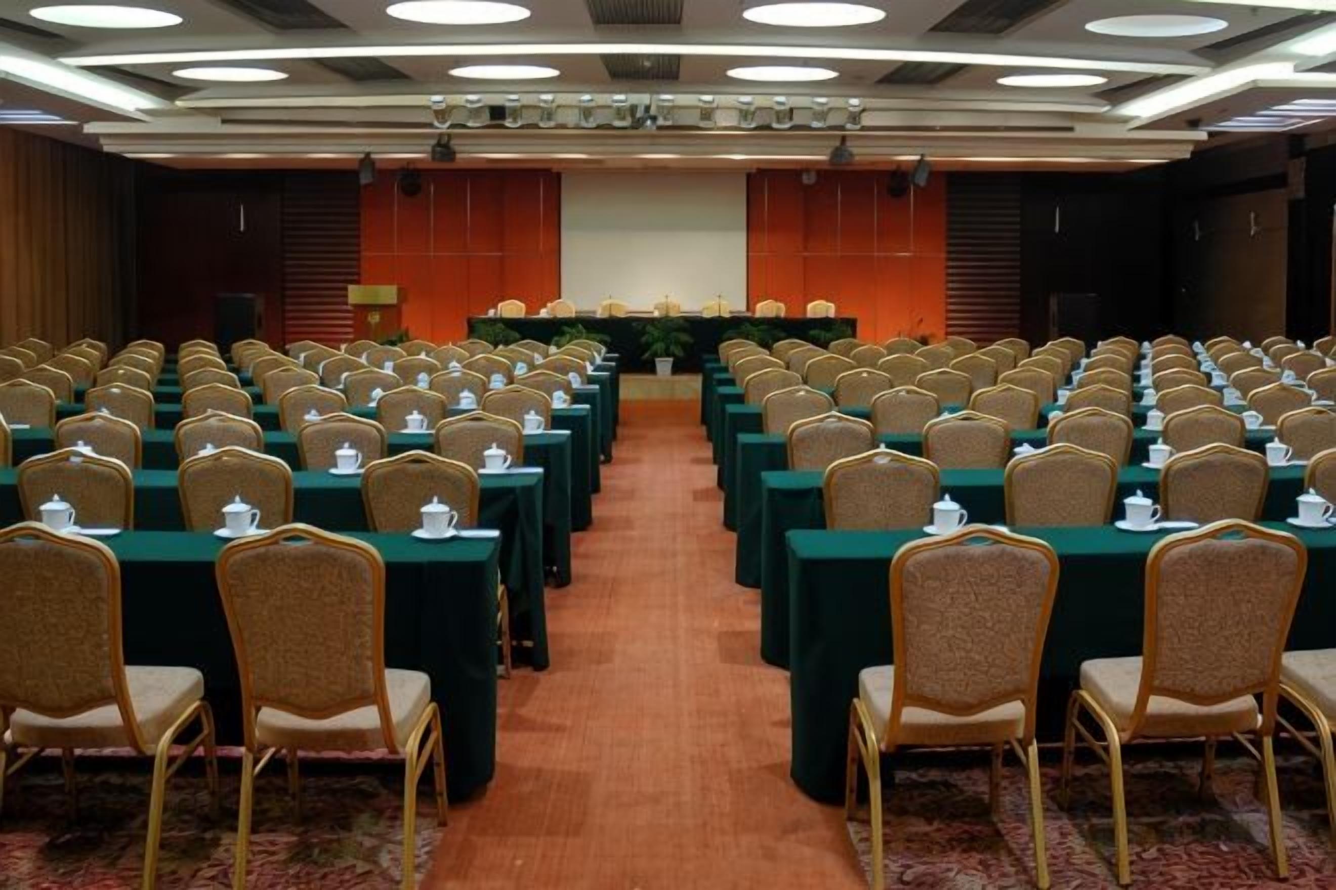 A conference room with rows of tables and chairs, a stage at the front, and a carpeted floor.
