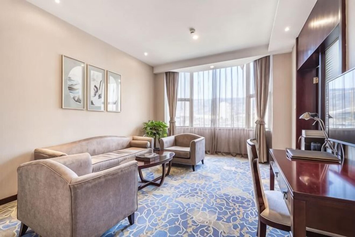 Habitación de hotel con sofá, sillón, mesa de comedor y un gran ventanal con cortinas.