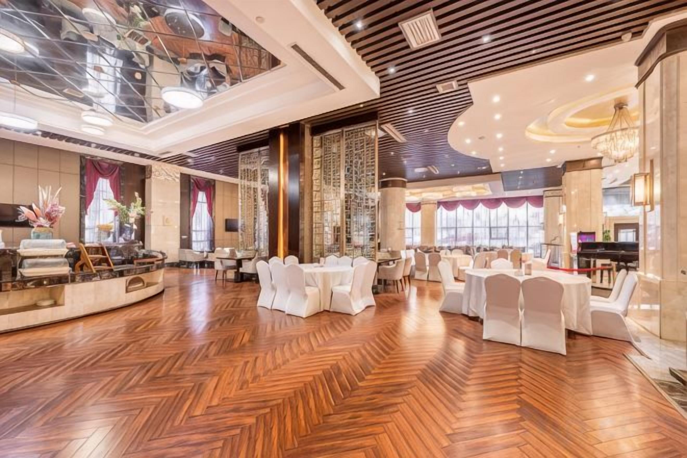 A spacious hotel lobby with a high ceiling, polished wooden flooring, and a central seating area with round tables and white chairs.
