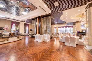 A spacious hotel lobby with a high ceiling, polished wooden flooring, and a central seating area with round tables and white chairs.