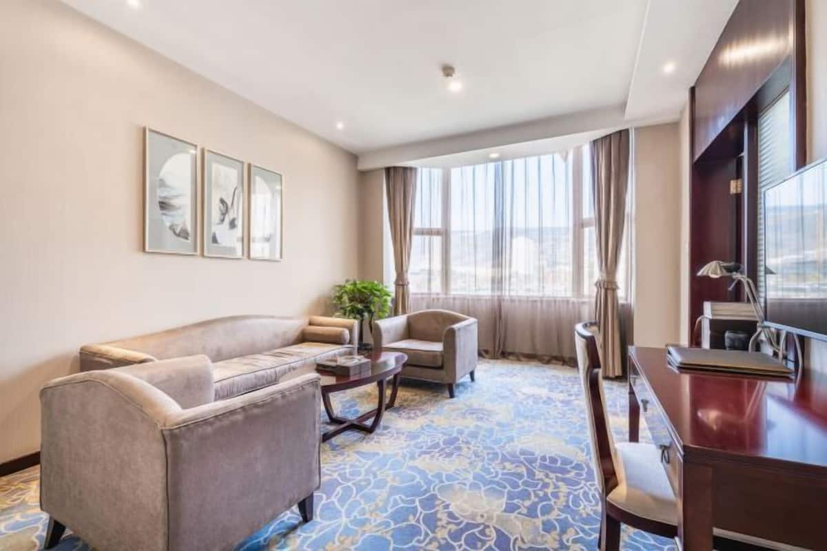 A hotel room with a sofa, armchairs, a coffee table, a TV, and a large window with curtains.