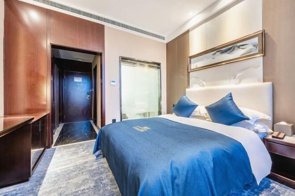 A hotel room with a large bed, a wooden wardrobe, a desk, and a glass door leading to a bathroom.