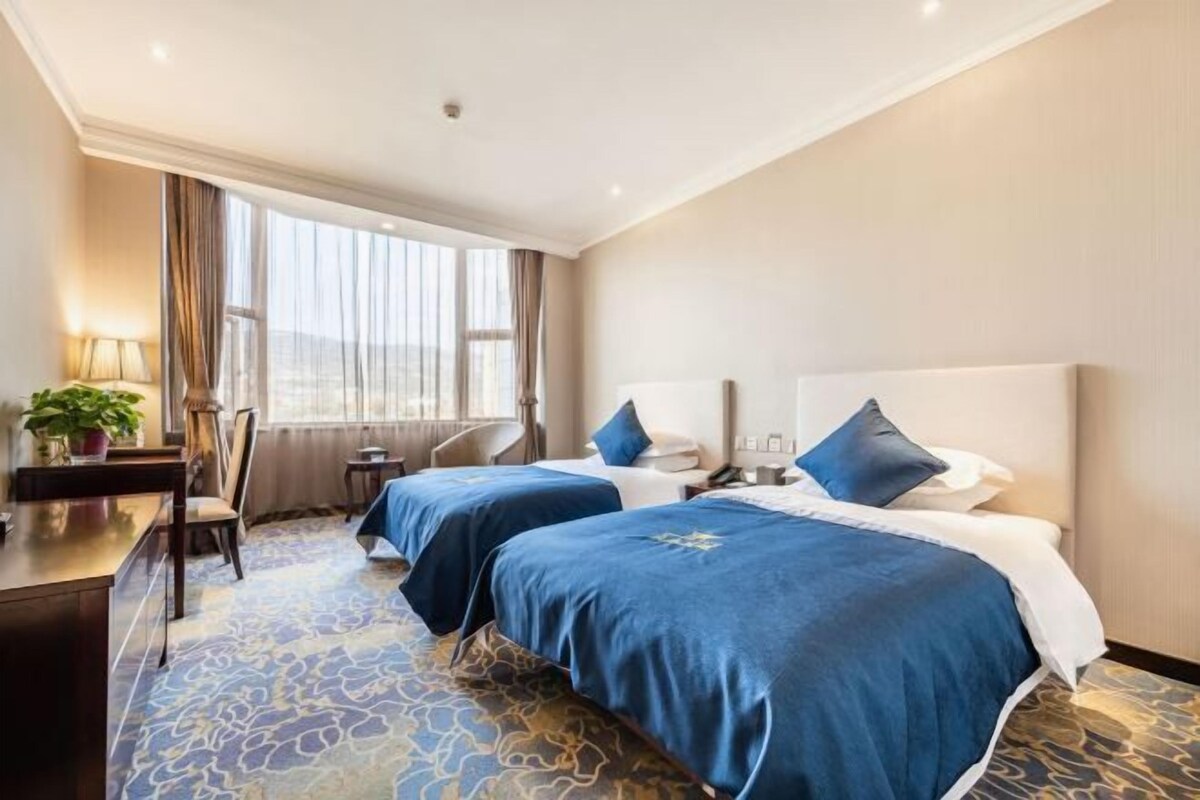 A hotel room with two beds, a desk, a chair, a lamp, and a view of the city.