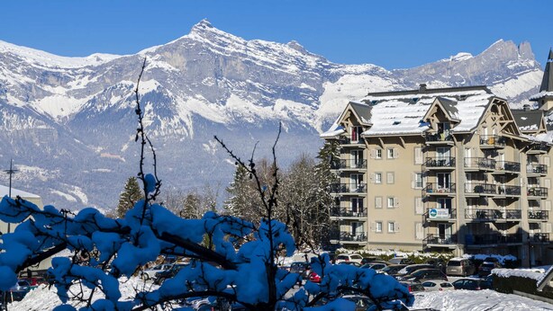 Ein Hotel mit mehreren Balkonen, einem verschneiten Berg im Hintergrund und einem Parkplatz mit Autos.