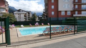 Ein Swimmingpool mit Liegestühlen, umgeben von einem grünen Zaun, mit Wohngebäuden im Hintergrund.