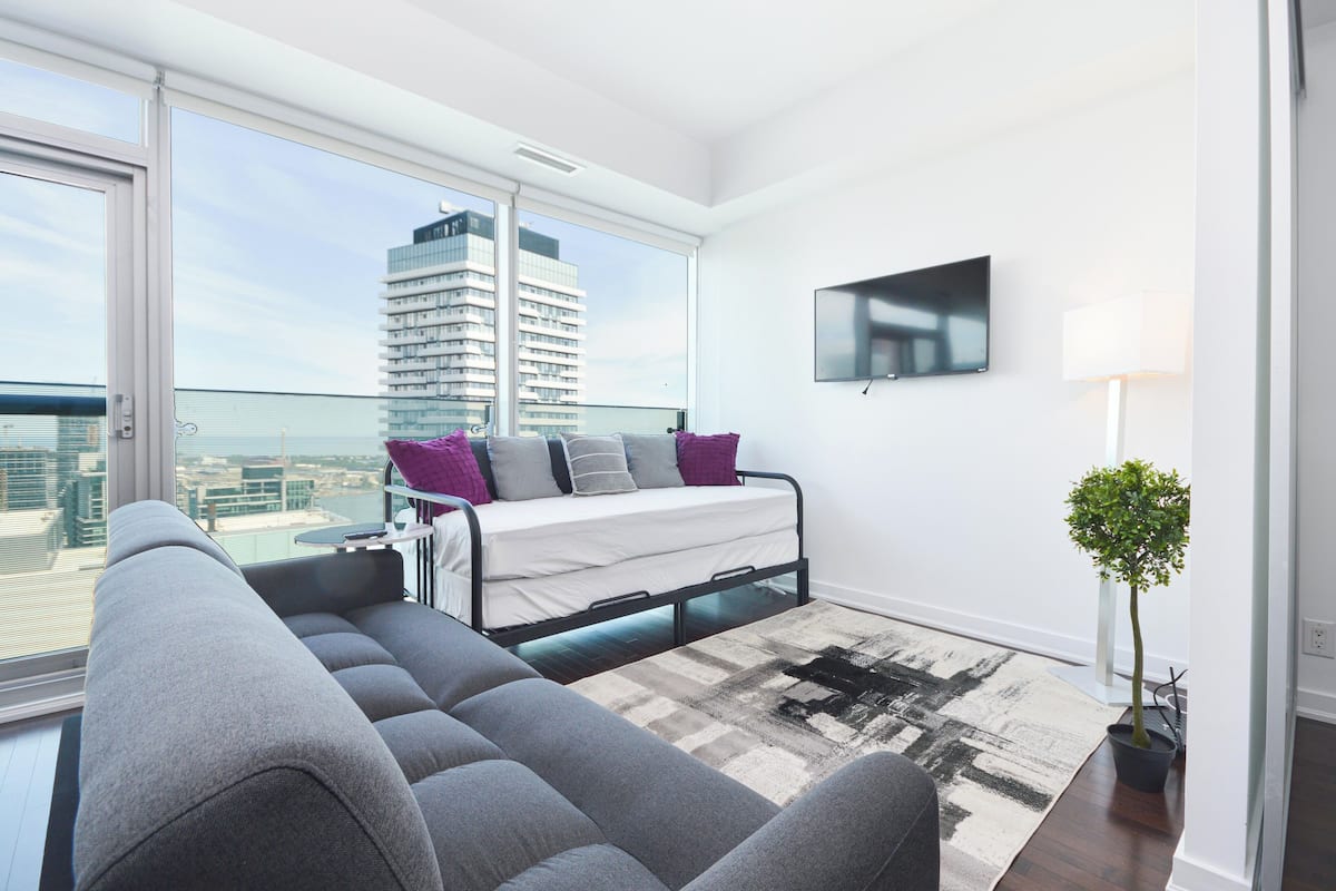 A modern bedroom with a bed, a grey sofa, a TV, and a view of a cityscape through large windows.