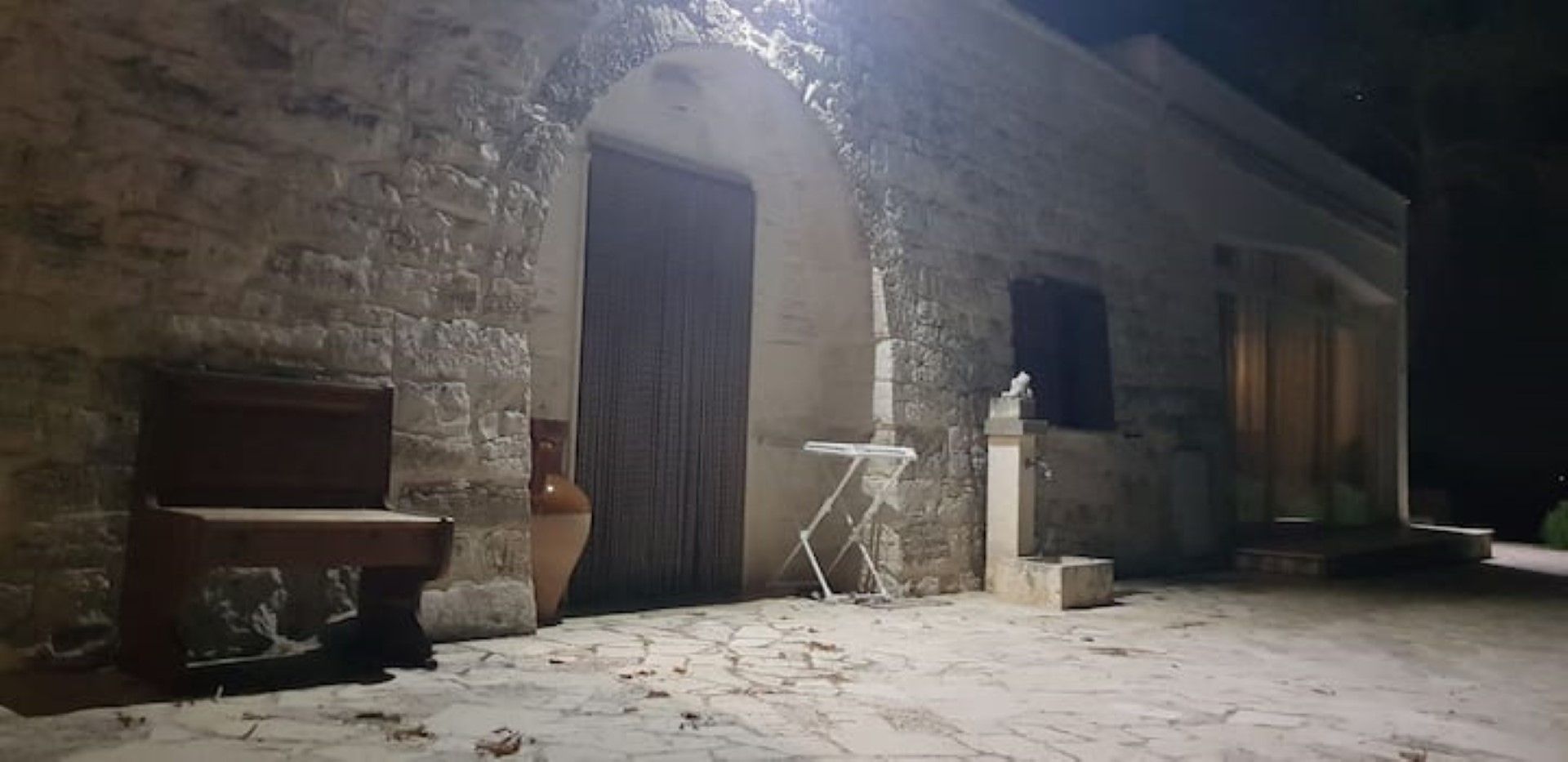 Un bâtiment en pierre avec une grande porte, un banc, une petite table et une statue.