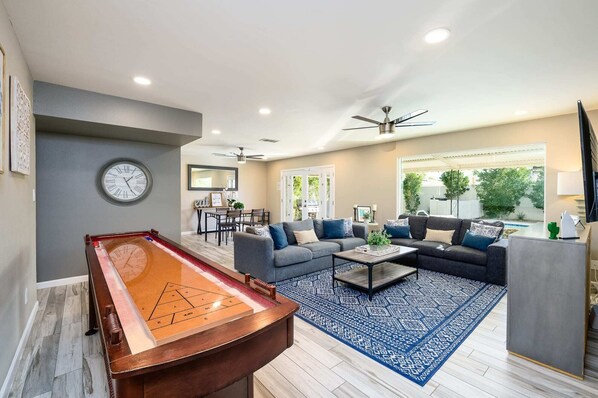 A living room with a pool table, grey sofas, and a dining area with a mirror and framed artwork.