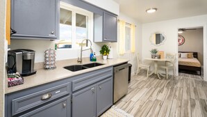 A compact kitchen with grey cabinets, a sink, and a dishwasher. There is a dining area with a table and chairs, and a bed in the adjacent room. - Old Town 4BR Gem | Pool, Firepit, WiFi, & Games! (Scottsdale)