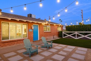 Une maison en briques avec une porte bleue, deux fauteuils Adirondack et des guirlandes lumineuses.