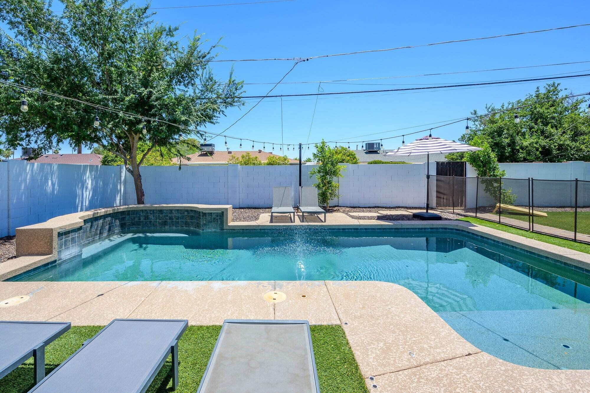 A rectangular swimming pool with a tiled edge, surrounded by a white wall and a fence. There are two lounge chairs and a small tree in the background.