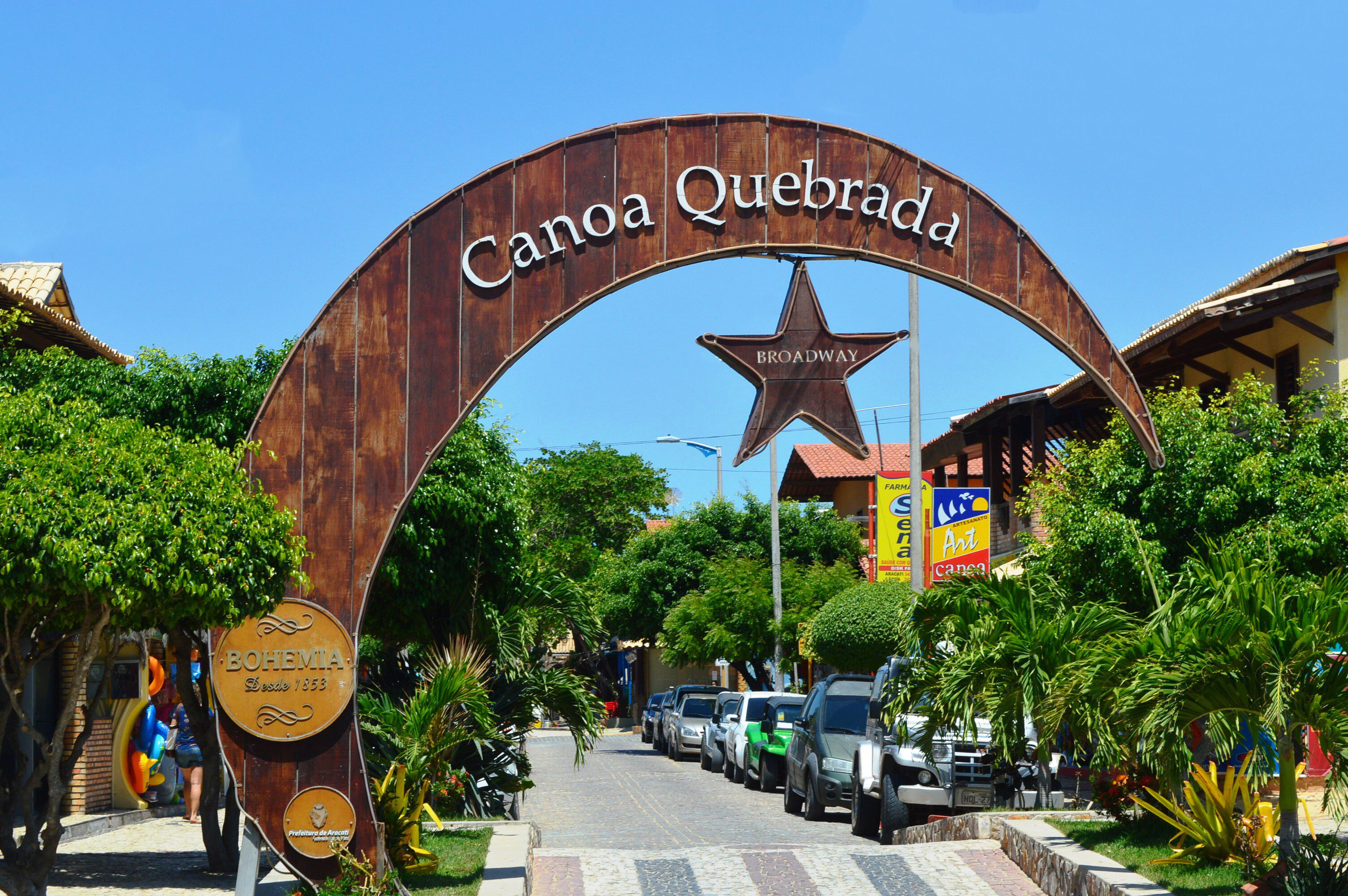 A large archway with "Canoa Quebrada" and "BOHEMIA" signs, a star, and a street with parked cars and trees.