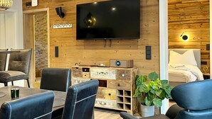 A modern living room with a flat-screen TV mounted on a wooden wall, a seating area with leather chairs, a small wooden side table, and a potted plant. - App. 5 - wohlB2 (3 Zi, 2 adult + 2 kids) (Braunlage)