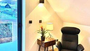 Una habitación moderna con un ventanal grande, una silla de oficina negra, una mesita de madera con una lámpara y una planta, y vistas a un paisaje nevado.