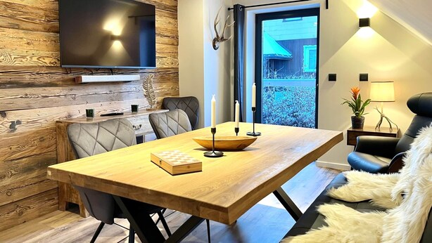 Un moderno comedor con una mesa de madera, sillas con cojines, un televisor de pantalla plana montado en una pared de madera y vistas al exterior a través de un ventanal.