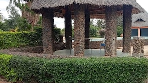 Un pabellón con techo de paja y pilares de piedra, una piscina y un edificio al fondo.