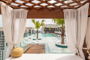A rooftop pool area with a wooden deck, palm trees, and a view of the cityscape.