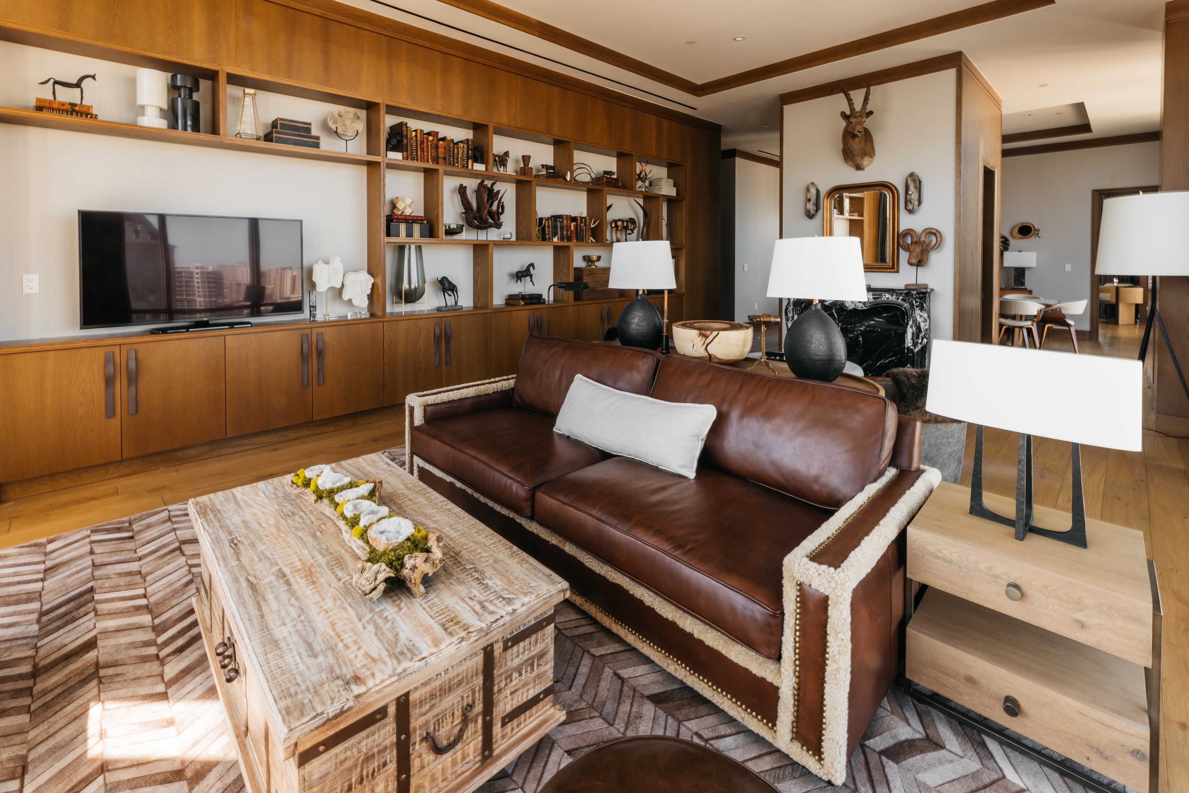 A living room with a brown leather sofa, wooden shelves with decorative items, a television, and a fireplace with two lamps.