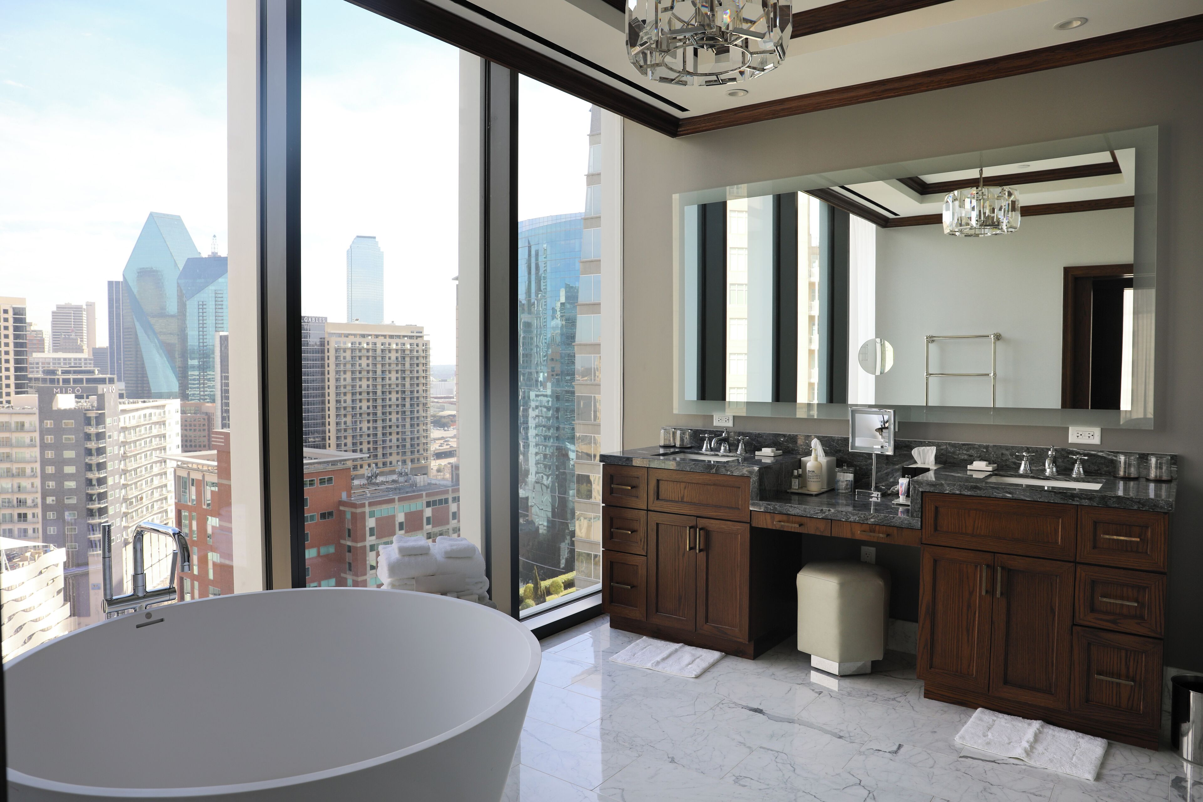 A modern bathroom with a large bathtub, dual sinks, and a large mirror. It features a city view through large windows.