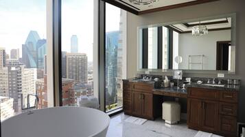 A modern bathroom with a large bathtub, dual sinks, and a large mirror. It features a city view through large windows.
