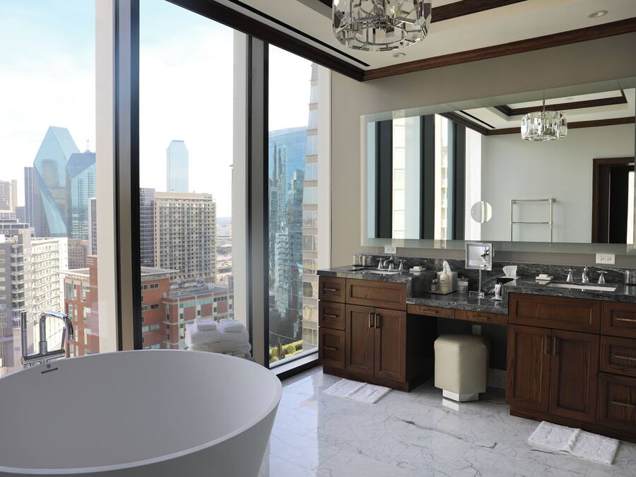 A modern bathroom with a large bathtub, dual sinks, and a large mirror. It features a city view through large windows.