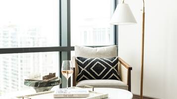 Quarto moderno com uma janela grande, uma mesa redonda com um livro e um copo de vinho, uma cadeira com almofada estampada e um abajur.