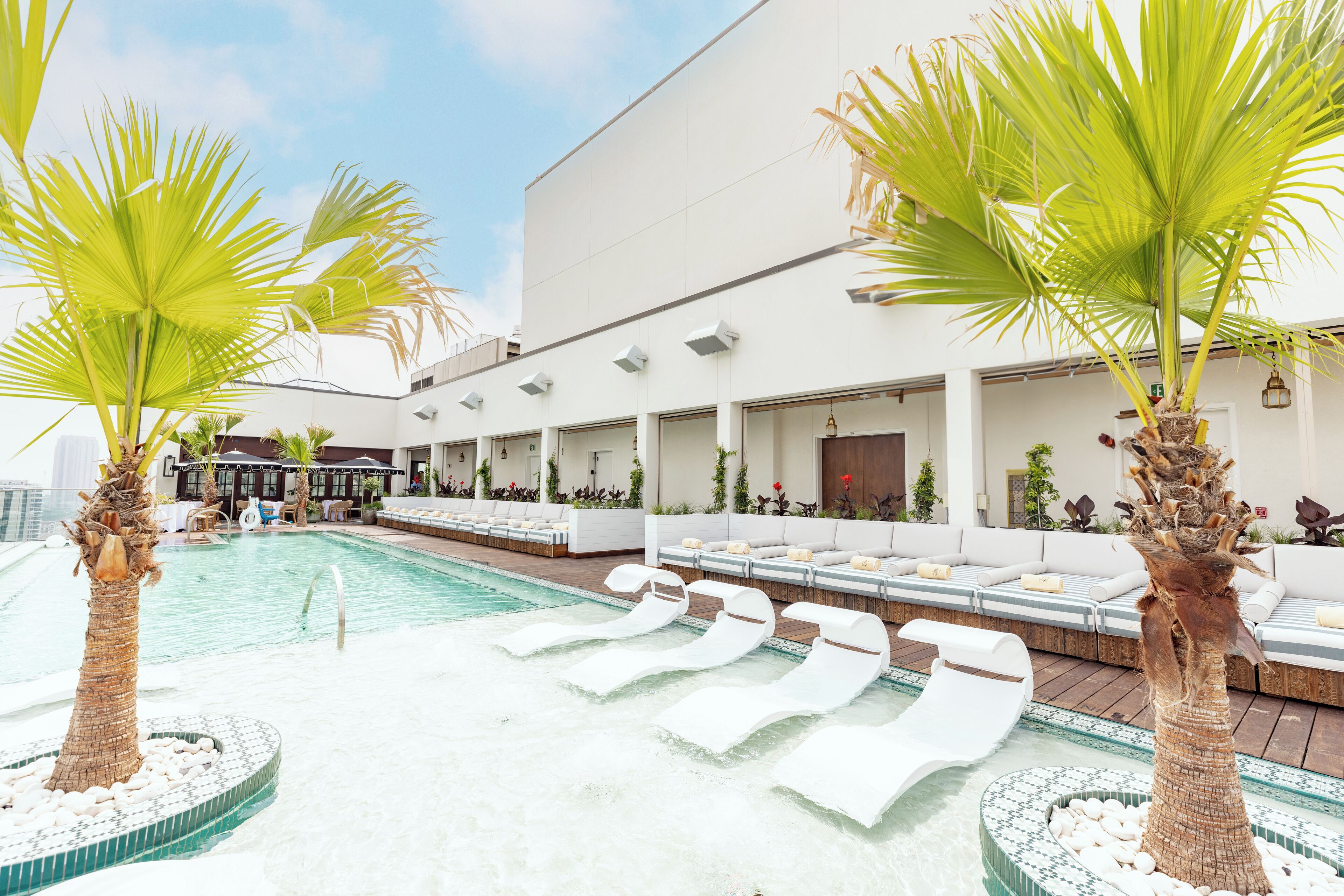 A pool area with lounge chairs, palm trees, and a building with outdoor seating.