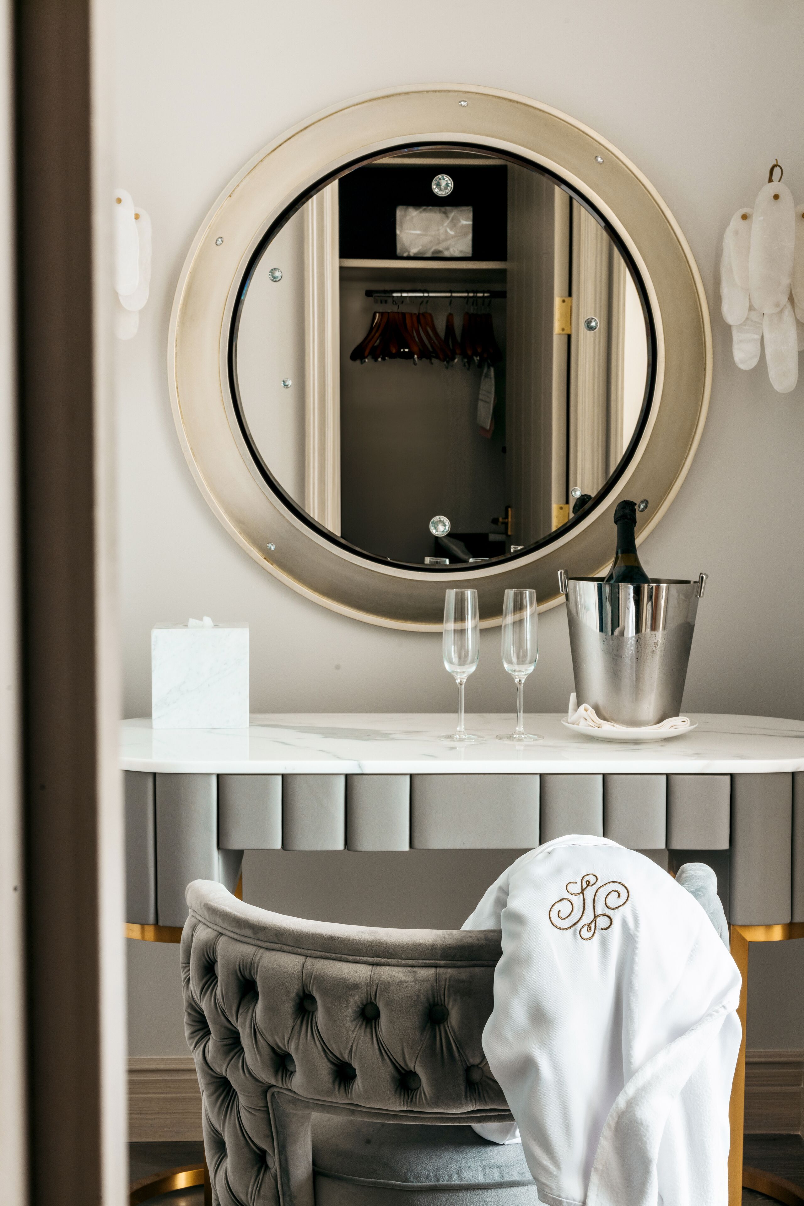 A bathroom with a round mirror, a vanity with a marble countertop, a champagne bucket, and a tufted chair.