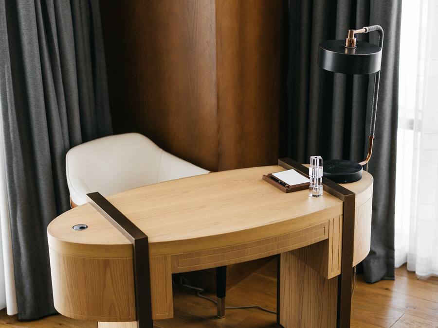 Habitació moderna amb escriptori de fusta, cadira blanca, làmpada de peu negra i otomana de cuir.